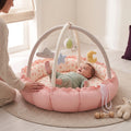 Baby looking up at the hanging toys in the pink play gym nest, with a parent close by