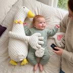 Baby lying on their back beside the white duck patting pillow, with a parent holding the remote